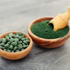 Due ciotole in legno: una con compresse verdi e l'altra con polvere di **pianta alga spirulina**, con un cucchiaino di legno.