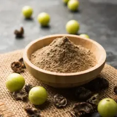 Polvere di triphala in una ciotola di legno, con accanto frutti di amla e parti essiccate. Il triphala a cosa serve? Aiuta la digestione e il benessere generale.