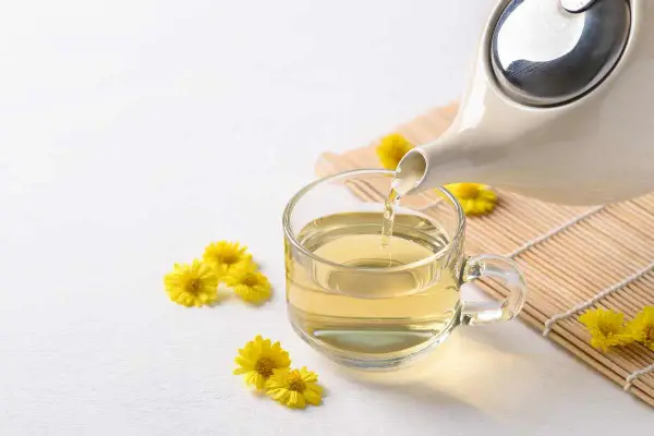 Tè giallo dorato versato da una teiera in una tazza trasparente, circondato da piccoli crisantemi fiori gialli.